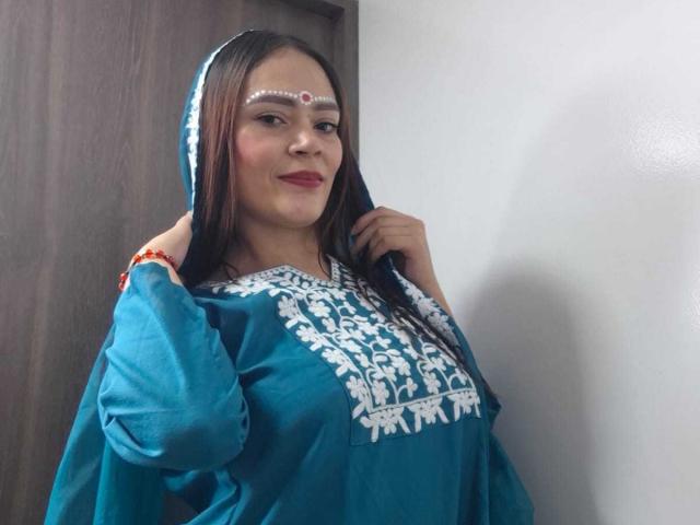 ShantalaKrisha stands in a room, wearing a blue traditional dress with white floral embroidery on the chest. She has long dark hair, a red lipstick, and a white decorative forehead piece. Her right hand pulls back the dress's hood, revealing a red bracelet on her wrist. The dress's sleeves are puffed, and the fabric looks slightly loose. She smiles slightly, looking at the camera. The background is a plain white wall and a wooden door.