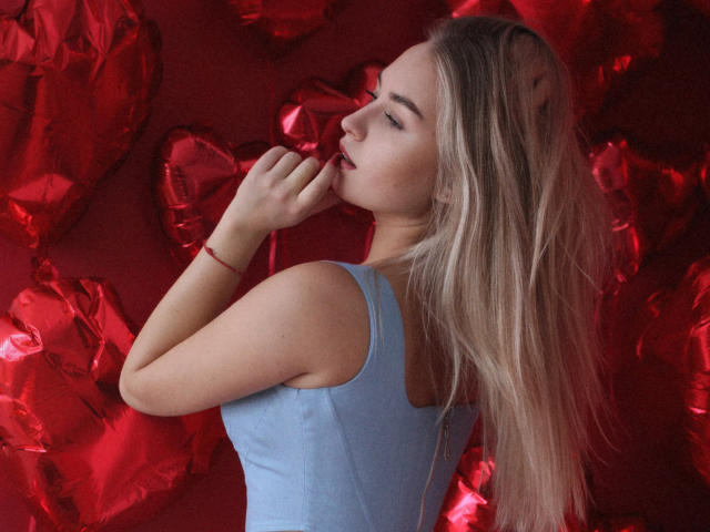 MargoWard with long blonde hair, blue tank top, red bracelet, fingers near mouth, red heart-shaped balloons, soft expression, slight smile, looking to the side, gentle light.