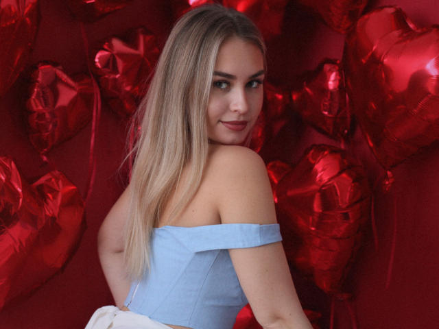 MargoWard with long blonde hair, blue off-shoulder top, smiling at camera, red heart-shaped balloons in background, showing her back and looking over shoulder, cute and sexy.