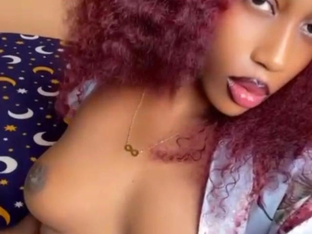 A slim-siren-hot with red curly hair, gold necklace, and pink lipstick, sticking out her tongue, exposing her small, dark-nipped tits. Her eyes are half-open, looking sideways.