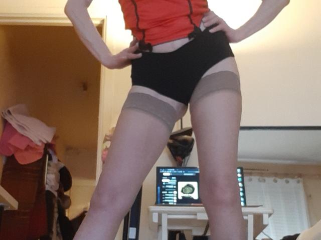 IssahneMay stands with hands on hips, wearing black panties, sheer thigh-high stockings, and an orange top, showing her bare thighs and a slightly visible pussy.