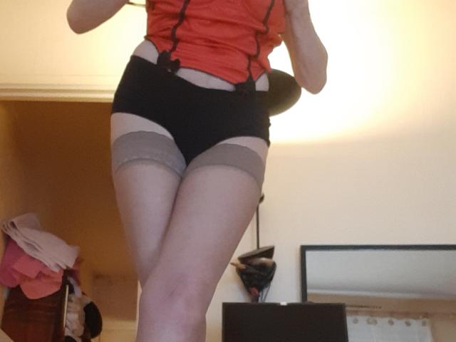 IssahneMay with pale skin, red top, black shorts, and gray thigh-highs, showing her ass and thighs, standing in a room with a slightly blurred background.