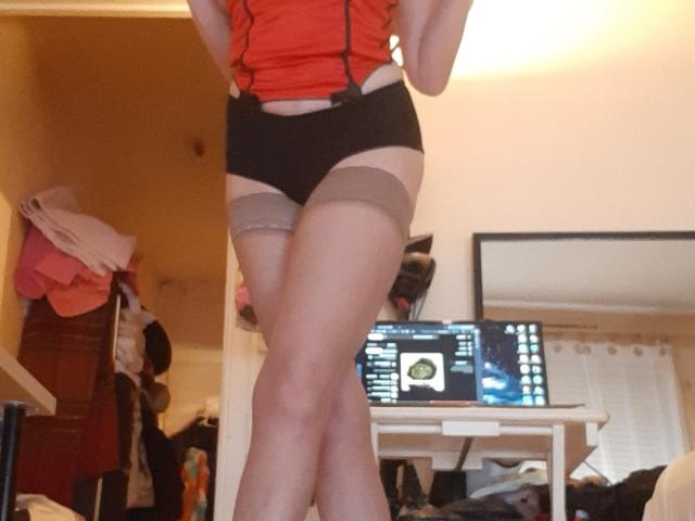 IssahneMay in red top, black shorts, and beige thigh-highs, standing with legs crossed, showing her ass and thighs, in a messy room with a laptop on a table.