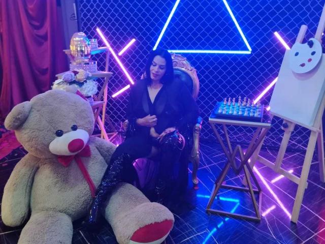 LizaRussell with black dress and shiny leggings, sitting on a chair, next to a giant teddy bear and neon lights, in a room with a chess set and painting.
