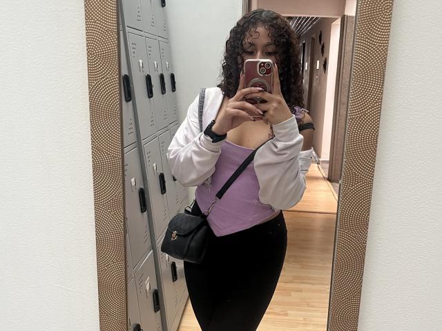 SofiaPassionne, con cabello rizado, blusa morada, chaqueta blanca, leggings negros, bolso negro, sosteniendo un teléfono, mostrando sus senos, de pie frente a un espejo, vestuario..