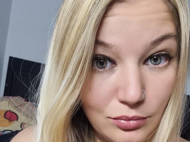 AlmaDeAngel69 has blonde hair, blue eyes, a nose ring, and pink lips. Her face is centered in the image, with her gaze directly at the camera.