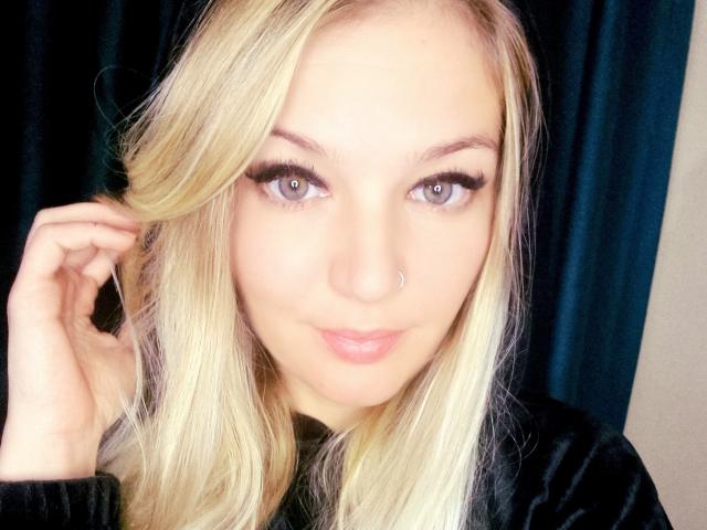 AlmaDeAngel69 has blonde hair, blue eyes, and a nose ring, with long eyelashes and a slight smile. She's wearing a black velvet shirt, touching her hair.