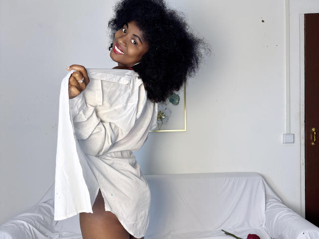 BeauteNoire with dark skin and a big afro, wearing a white shirt, holding it open to show her ass, smiling at the camera. White sofa and door in background.