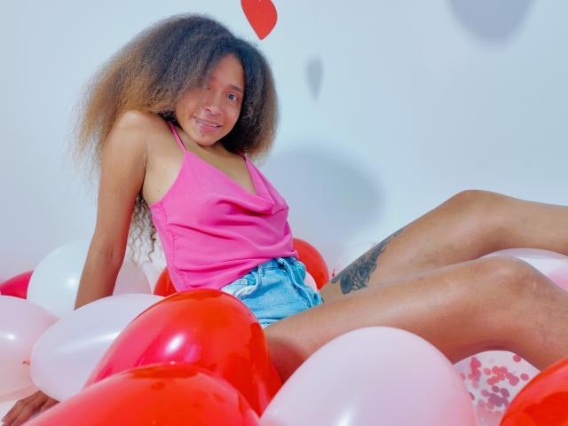 SweetSexx with curly brown hair, pink tank top, and denim shorts, sitting in red and white balloons, showing a tattoo on her right thigh and smiling.