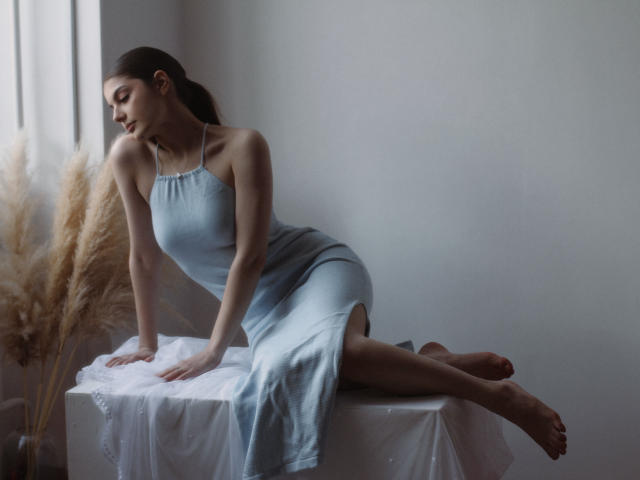 StellaLewin with small tits, wearing a light blue dress, leans on a white-covered table, legs stretched, puffy reed plant on left, soft natural light, calm expression.