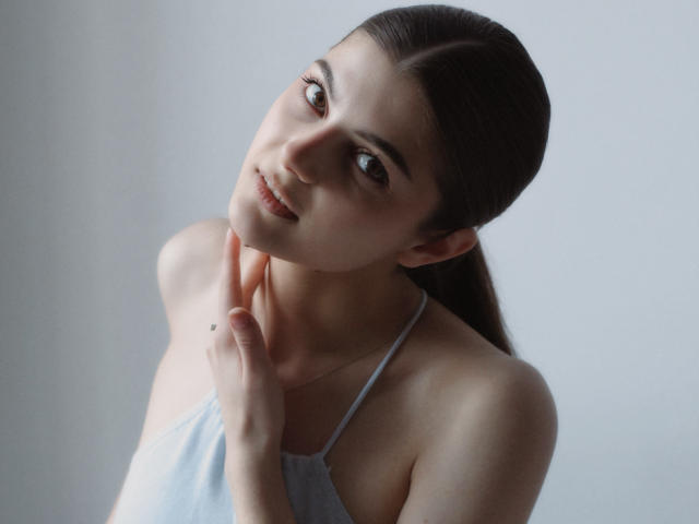 StellaLewin with light skin and brown hair, wearing a white halter top, gently touching her neck, smiling softly, brown eyes, slender, cute, and subtle makeup.