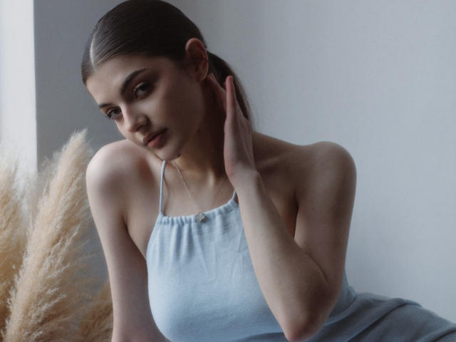 StellaLewin with puffy brown hair, wearing a light blue halter top, touching her neck, showing her small tits, and looking down with a slight smile. Puffy grass on left.
