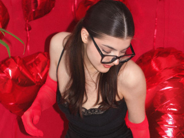 StellaLewin with red gloves, black lingerie, and glasses, looking down, showing her tits, in a red heart-themed background.