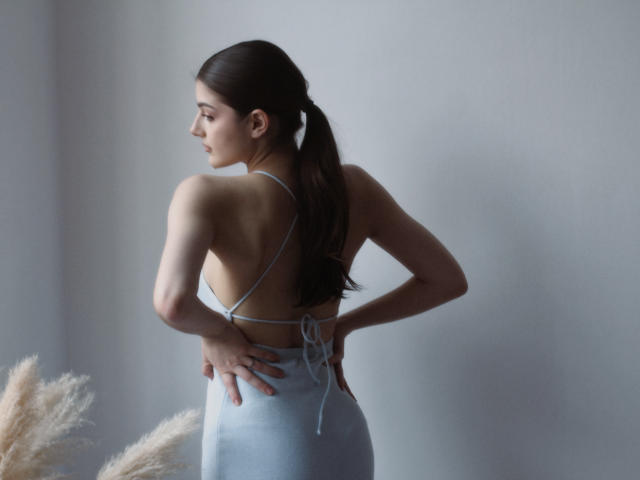 StellaLewin, back to camera, hands on hips, wearing a halter blue dress, showing her ass, long brown ponytail, light skin, soft gray background, puffy white grass in foreground.