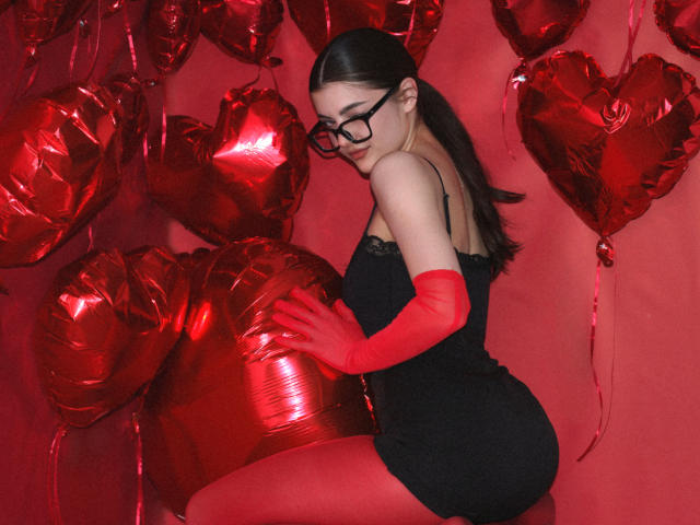 StellaLewin with glasses, black dress, red gloves, and red tights, kneeling, touching shiny red heart balloons, red background, long dark hair, looking over shoulder.
