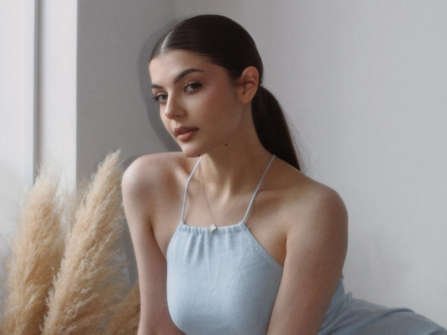 StellaLewin with long dark hair, wearing a light blue halter top, showing small tits and a slim figure, standing near puffy grass, looking to the side with a subtle smile.