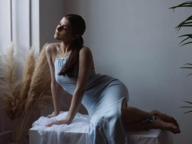 StellaLewin with her ass up, wearing a light blue dress, leaning on a white table, puffy tits, brown hair in a ponytail, eyes closed, soft light, pampas grass.