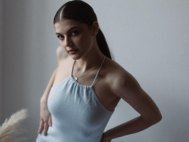 StellaLewin with slicked-back brown hair, wearing a light blue halter top, showing small tits and a slim, pale body, standing with hands on hips, looking at camera.