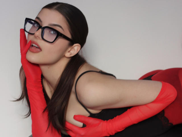 StellaLewin with glasses, red gloves, black top, and red pants, leaning forward, hands on face, tongue slightly out, brown hair, white background, sexy, attractive.