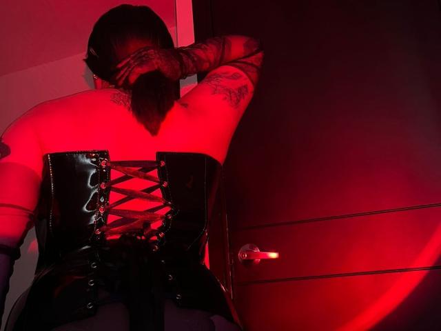 Jesskhy with tattooed arms, black leather corset, laced up, red light, back to camera, ponytail, standing by a door, red glow, black leather, visible metal handle.