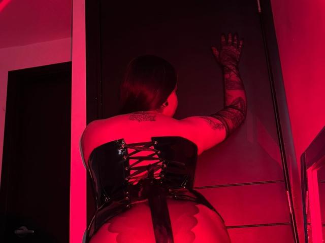 Jesskhy's back is to the camera, wearing a black, shiny, lace-up corset, exposing her ass, and raising a tattooed arm against a red-lit door.