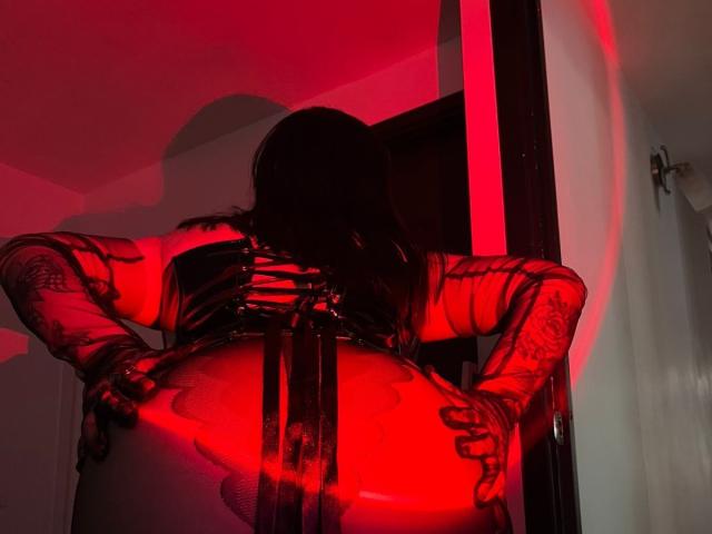 Jesskhy with tattoos, back to camera, wearing black latex corset, red light, hands on large butt, long dark hair, red light casting shadow, in dimly lit room.