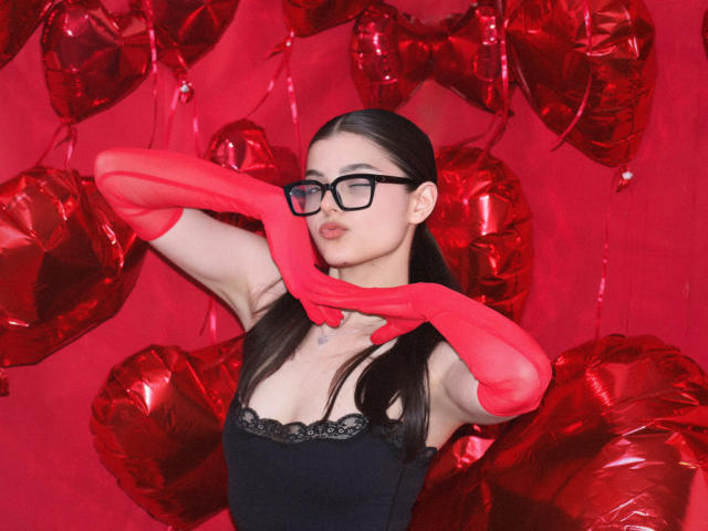StellaLewin with red gloves, black lace top, and glasses, holding gloves up, showing her tits, against a red heart balloon background. She's a cute, nerdy, sexy bitch.