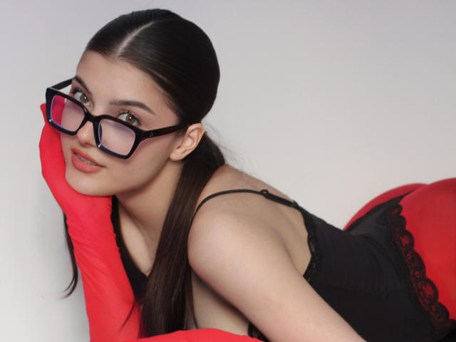 StellaLewin with black glasses, red gloves, and black lace top, leaning on her hand, showing off her ass. Long brown hair, pale skin, and a sexy smile.