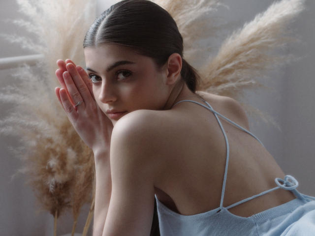 StellaLewin with pouty face, hands near mouth, wearing a light blue spaghetti-strap top, brown puffy hair in ponytail, pale skin, soft light, fluffy beige pampas grass in background.