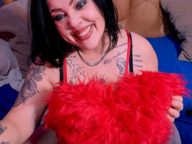 SatisfyMyDesires with black hair, tattoos, and red lipstick, holding a red, fluffy heart, smiling, wearing a red and black strap top, sitting on a blue bed.