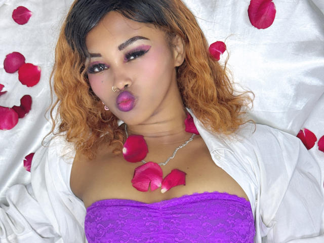 LaCoquineX with curly orange hair, purple lace top, white shirt, rose petals, and nose ring, pouting with pink lipstick, silver necklace, and pink eye makeup.