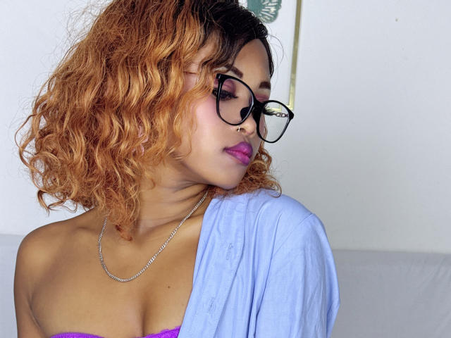 LaCoquineX with curly, orange-brown hair, wearing glasses, a blue shirt, and a purple bra, has pink lipstick and a nose ring, looking to the side, with a silver necklace.