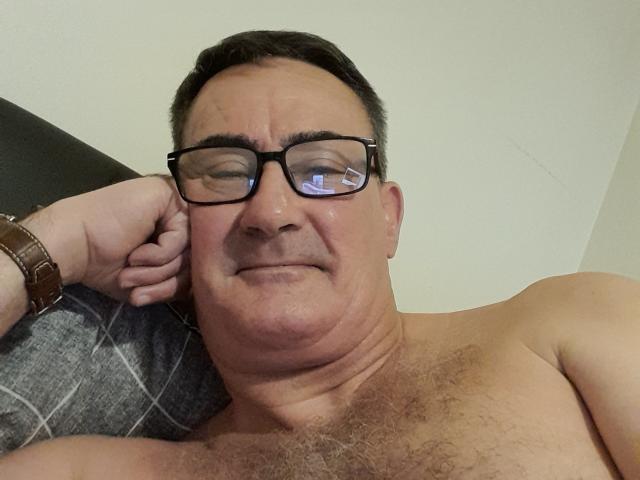 Shirtless StillGold with glasses, brown watch, and short dark hair, has a slight smile, hairy chest, and is leaning on a gray pillow.