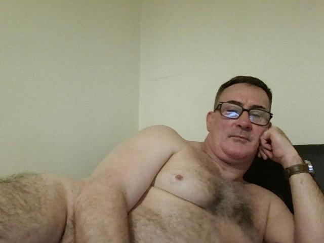 StillGold is a chubby, naked man with glasses, lying on a bed, chest hairy, right arm raised, wrist bracelet, looking slightly annoyed, short brown hair, pale skin.