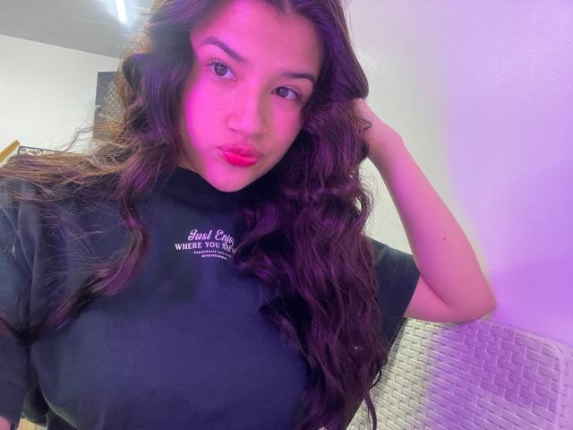 Iris Blair, met lang, golvend bruin haar, roze-paarse highlights en rode lippenstift, draagt een zwart T-shirt, frunselt met haar volle, glanzende roze lippen en heeft haar hand in haar haar..