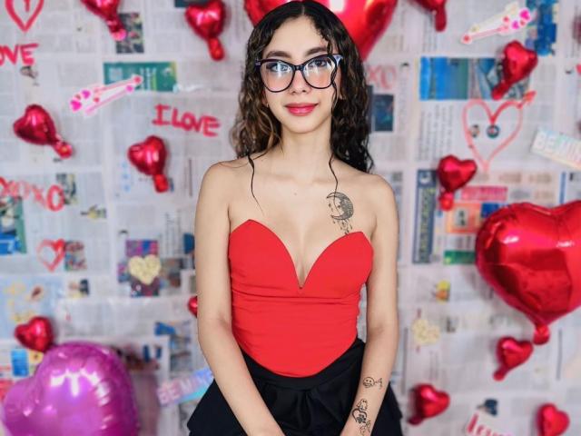 HotAndWildy with curly brown hair, glasses, red strapless top showing small tits, black skirt, tattoos, smiling, standing in front of red heart balloons and newspaper background.