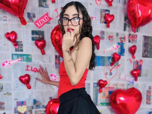 HotAndWildy with curly dark hair, red tank top, black skirt, glasses, and tattoo on hand, touching lips, surrounded by red heart balloons, looking cute and sexy.