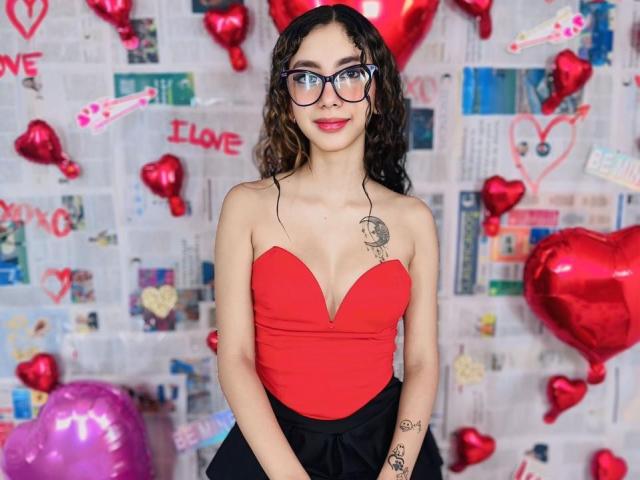 HotAndWildy with curly brown hair, glasses, red strapless top, black skirt, tattoos on right shoulder and left forearm, standing with red heart balloons and pink balloons.