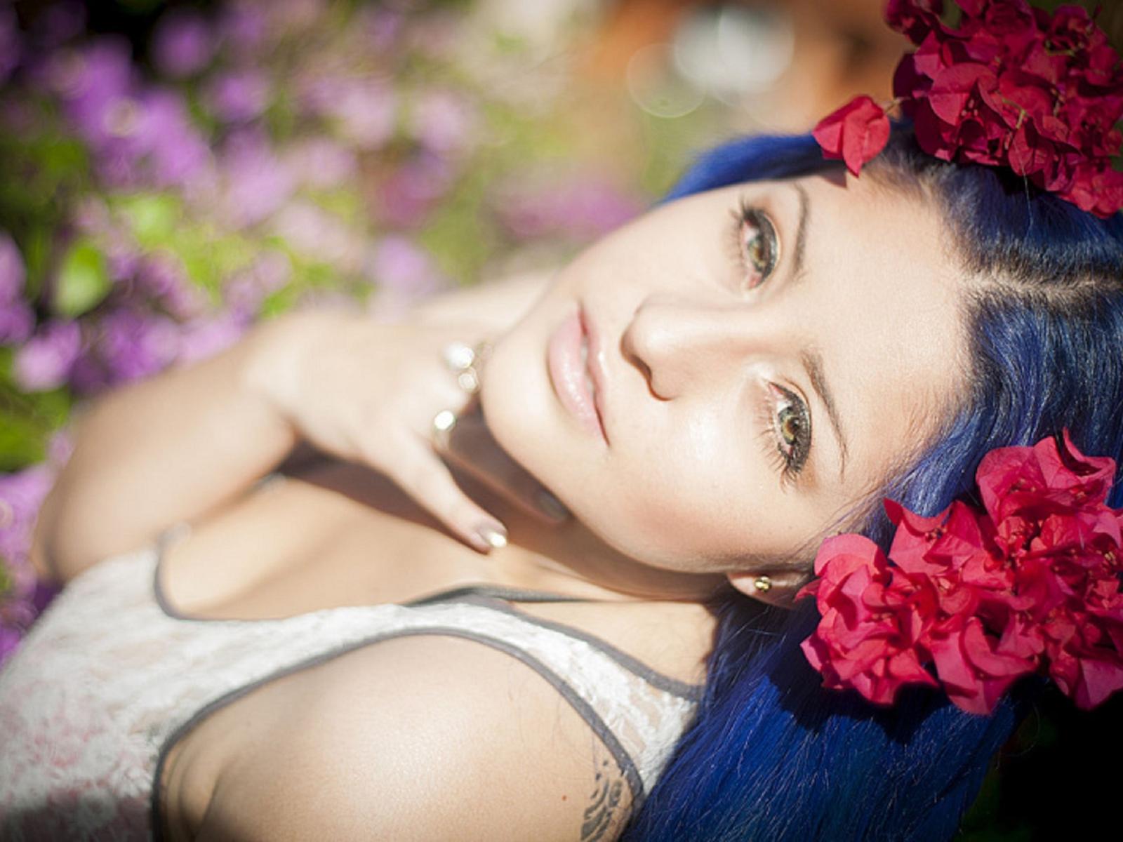 CarmelinaX with blue hair, wearing red flower headpiece, looking up with soft smile, pink lipstick, white lace top, light skin, green eyes, and hand near chin, sunlit.
