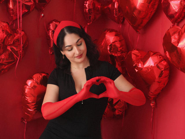 StefanaWinter with red gloves, black dress, and red headband, making a heart shape with her hands, surrounded by shiny red heart balloons, smiling gently.