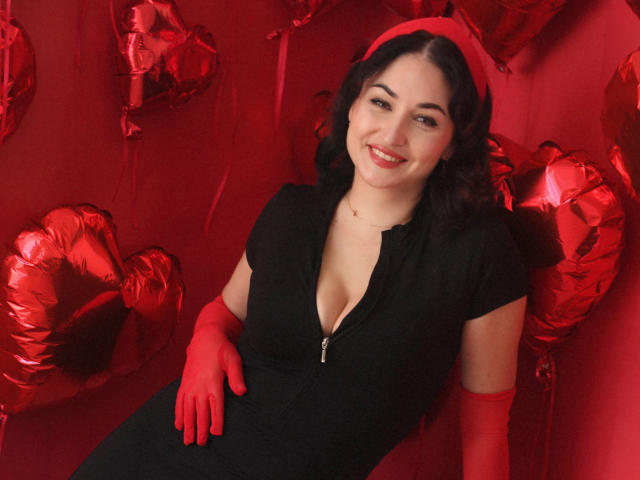 StefanaWinter with red gloves, black zip-up dress, showing her tits, smiling, red heart balloons, black wavy hair, red headband, red background, cute, sexy, playful.