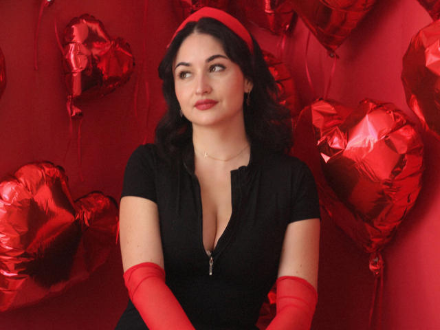 StefanaWinter with red heart balloons, wearing black zip-up dress, red gloves, and red headband, looking to the side with a slight smile, showing her tits.