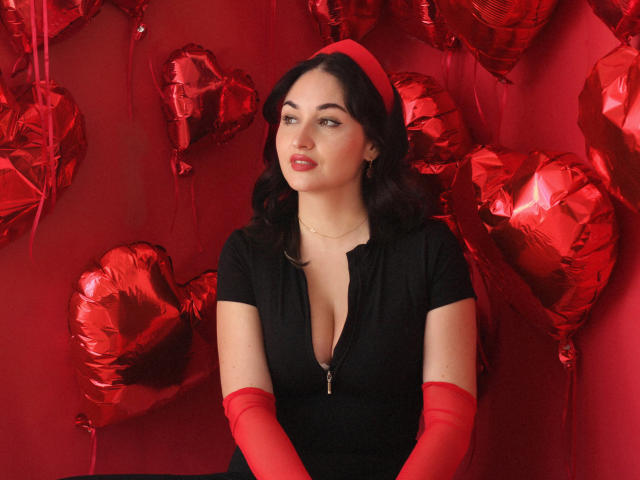 StefanaWinter with red heart balloons, black dress, deep cleavage, red gloves, and red headband, sitting against a red wall, looking slightly to the side, with a serious expression.