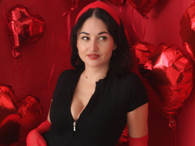 StefanaWinter with red lipstick, black zip-up top, showing her boobs, red heart balloons, and red gloves, smiling slightly, dark wavy hair, fair skin, red headband.