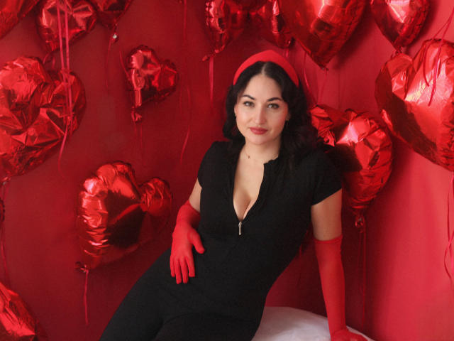 StefanaWinter with red gloves, black zip-up dress, and red headband, sitting against red heart balloons, showing a subtle smile, and her tits slightly visible through the dress.