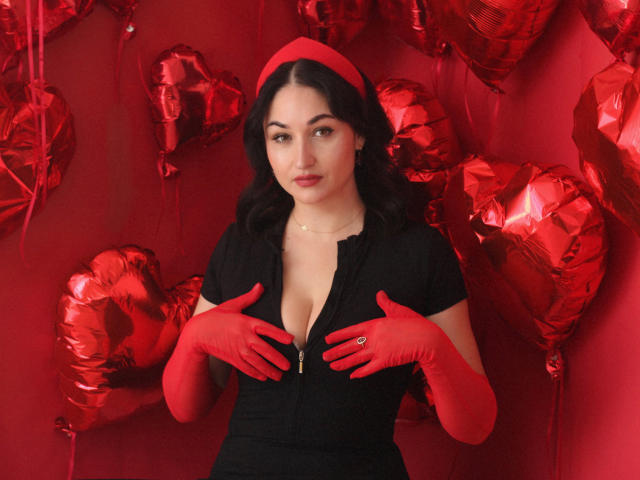 StefanaWinter with red gloves, black dress, and red headband, hands on her tits, surrounded by shiny red heart balloons. She has fair skin, dark wavy hair, and red lipstick.