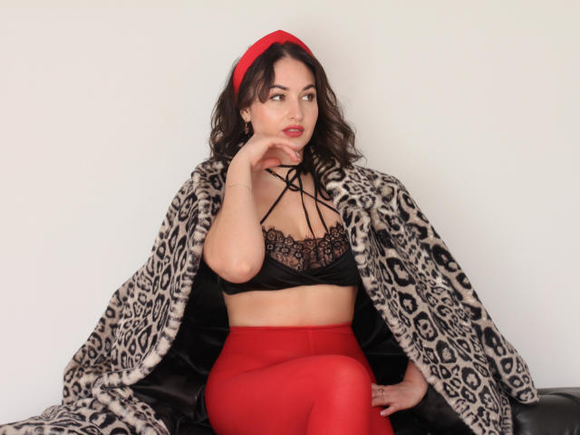 StefanaWinter with red headband, black lace top, and red pants, wearing a leopard fur coat, sitting on a black leather surface, hand on chin, looking to the side.