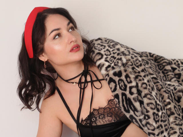 StefanaWinter with dark hair, red headband, black lace top, small tits, black ribbon choker, looking up, leopard fur blanket, red lipstick, gold earrings, white background.