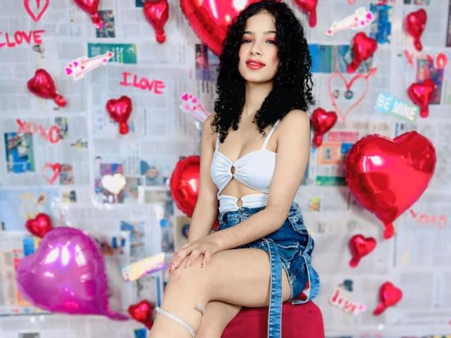 BellainLove with curly black hair, white bra with gold ring, and denim shorts, sitting on red chair, smiling, red heart balloons, pink balloon, white wall, sexy, cute.
