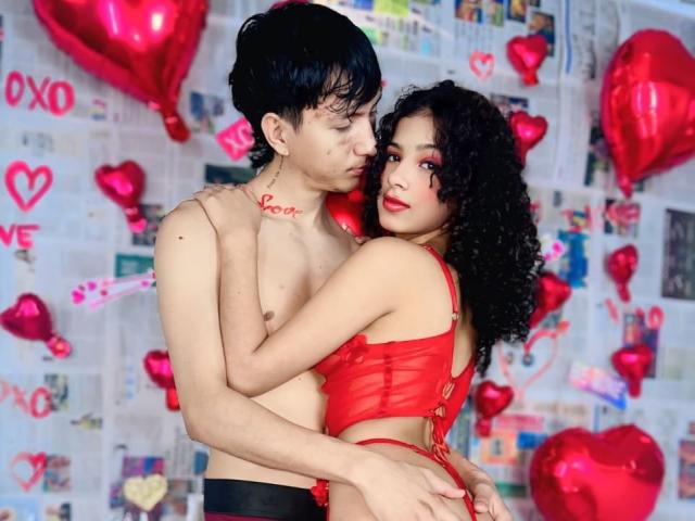 A photograph of BellainLove with a slim, Asian male and a curvy, Latina female. Both are topless; the male has short, black hair and "Love" tattooed on his chest, wearing black pants. The female has curly black hair, wears red lace lingerie, and looks at the camera with red lipstick. They are intimately embracing. Red heart-shaped balloons and "Love" graffiti decorate the background. The male's left hand is on her back, while her right hand holds his buttock. The composition emphasizes their physical connection and romantic theme.